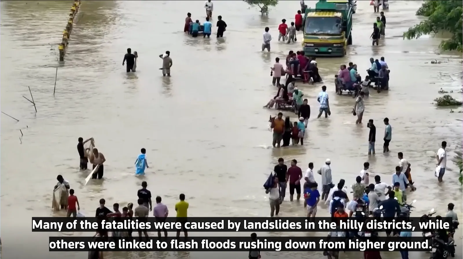 Dozens of people wade through a deeply flooded road beside a stranded truck